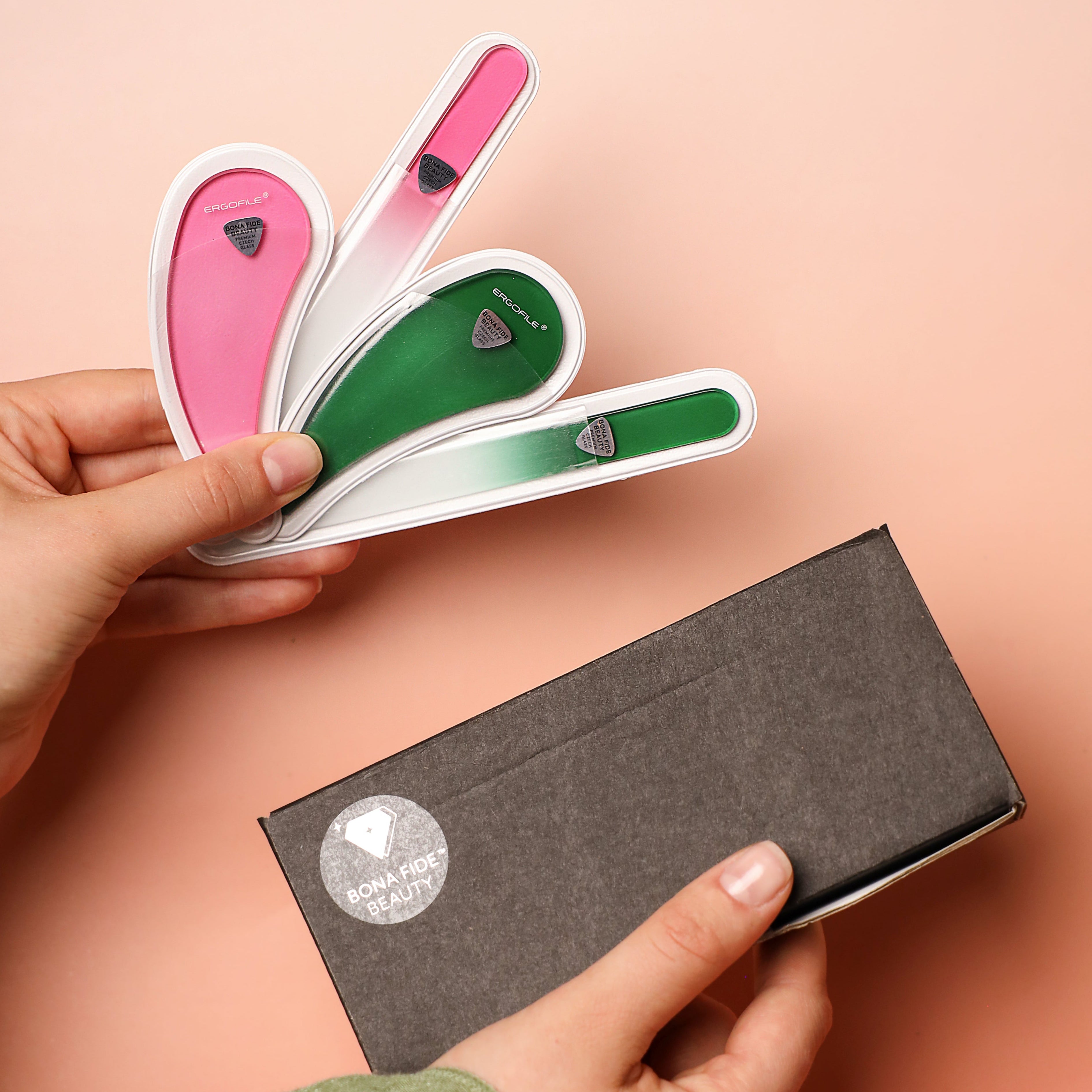 Hands holding a green and pink tear-dropped glass nail files that are more ergonomic to hold, with a black box and a peach background